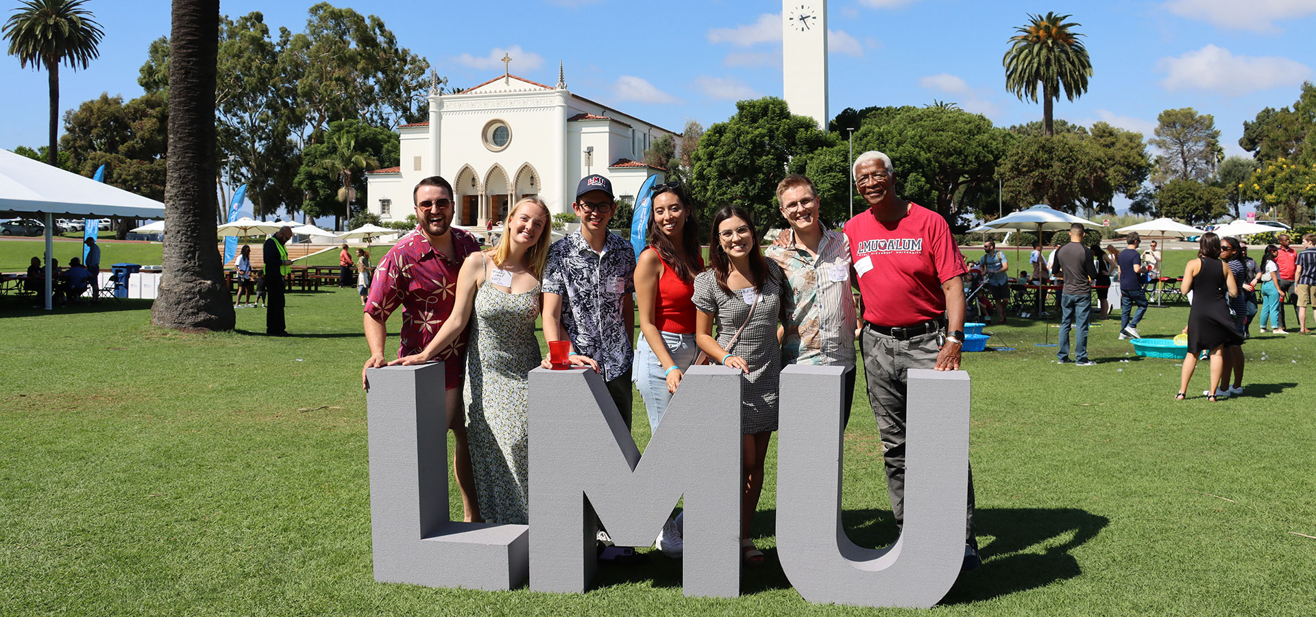 Several alumni standing behind large LMU block letters on Sunken Garden with Sacred Heart Chapel in the background