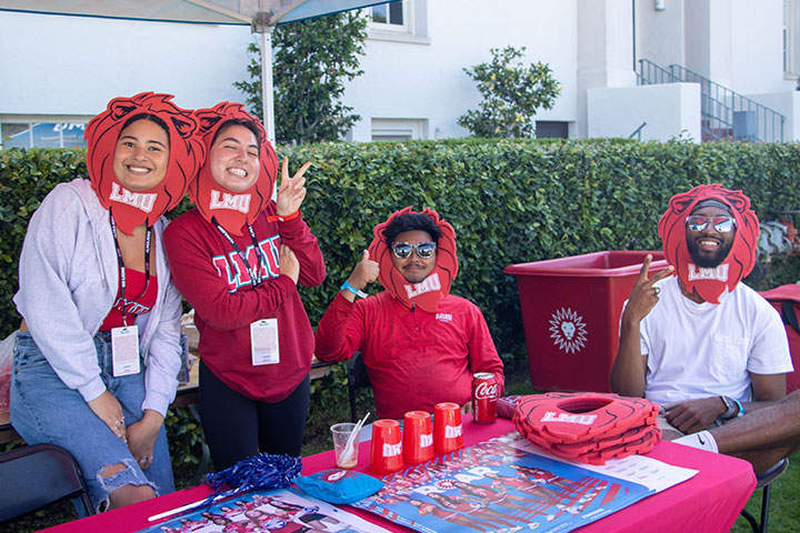 Four people smile wearing LMU gear and a lion's mane costume on their heads