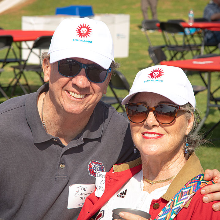 Two people in matching white LMU Alumni hats smile during LMU Alumni BBQ